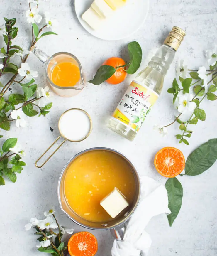 A saucepan filled with a thick bright orange liquid sits in the center of a white marbled surface, with a large square of pale yellow butter floating on top near the right side, wrapped in a white cloth knotted at the handle below. To the upper left is a small clear glass pitcher filled with the same orange liquid, while a brass measuring cup filled with white sugar rests just below it. At the top center, a white plate holds four pale yellow pats of butter. To the right of the plate is a clear glass bottle with a yellow label and golden cap showing orange blossoms. Two bright orange tangerines with green leaves attached, one whole and one halved exposing the juicy segmented inside, sit near the bottom and right side of the saucepan. White blossoming green leafy branches frame the image on the left and right edges. Photo taken with an iphone --ar 4:5 --v 7