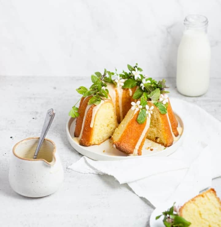 A round, yellow cake with a smooth texture is placed on a white plate, decorated on top with green leaves and small white flowers. The cake has three visible layers, each separated by a light brown drizzle running down the side. Part of the cake is sliced, showing the soft inside. Next to the cake on the white marbled surface is a small white jug with a spoon inside, holding more light brown drizzle. In the background is a clear glass bottle filled with milk. The setting includes a white cloth partly under the plate. Photo taken with an iphone --ar 4:5 --v 7