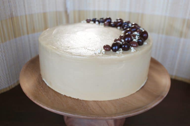 A smooth, round cake with a light beige cream frosting evenly covering its entire surface, sitting on a simple wooden cake stand. On top, there is a crescent-shaped decoration made of glossy dark brown round candies arranged along one edge, sprinkled with small white flakes around the candies. The background is a soft, light curtain with a subtle pattern, and the surface beneath the stand is a white marbled texture. photo taken with an iphone --ar 4:5 --v 7