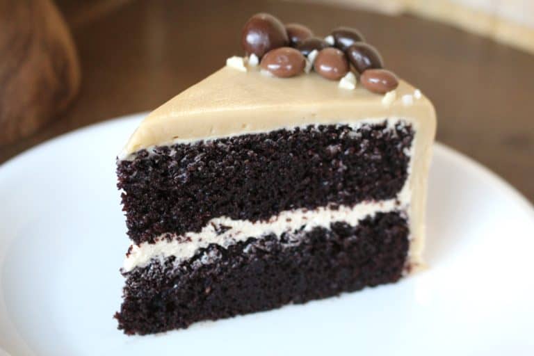 A slice of two-layer dark chocolate cake on a white plate, each layer thick and moist with a light cream filling between them. The cake is covered in a smooth light brown frosting on top and sides. On top of the slice, there are small round chocolate-covered nuts or candies scattered near the pointed edge. The plate rests on a white marbled surface. Photo taken with an iphone --ar 4:5 --v 7