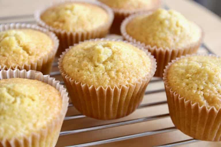 The image shows seven yellow cupcakes with slightly cracked tops in white paper liners arranged on a metal cooling rack over a white marbled surface. The cupcakes have a soft and fluffy texture with a light golden color. Each cupcake is round with a gently domed top, showcasing a crumbly, homemade look. The background is softly blurred, emphasizing the cupcakes in the center. Photo taken with an iphone --ar 4:5 --v 7