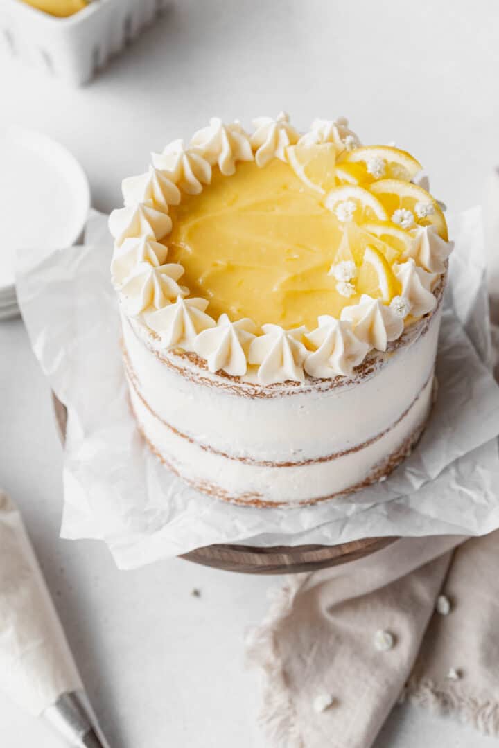 A two-layer cake sits on crumpled white parchment on a wooden round board, the cake layers are light tan with a smooth white frosting thinly covering the sides, showing the cake underneath. The top layer is covered with a glossy yellow lemon filling. Around the edge of the top, there are evenly spaced white frosting peaks, with a cluster of lemon slices and small white frosting flowers on one side. The whole scene is set on a white marbled surface with soft, neutral-colored cloth and small white decorations scattered nearby. Photo taken with an iphone --ar 4:5 --v 7