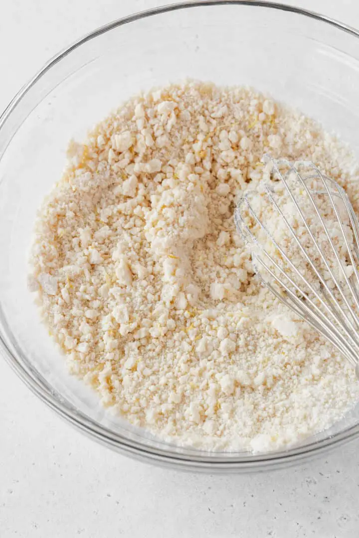 A clear glass bowl contains a mix of dry ingredients with a crumbly texture showing small, pale lumps spread evenly across the surface. On the right side inside the bowl lies a metal whisk with thin wires coated lightly by the mixture. The bowl is placed on a white marbled surface. photo taken with an iphone --ar 4:5 --v 7