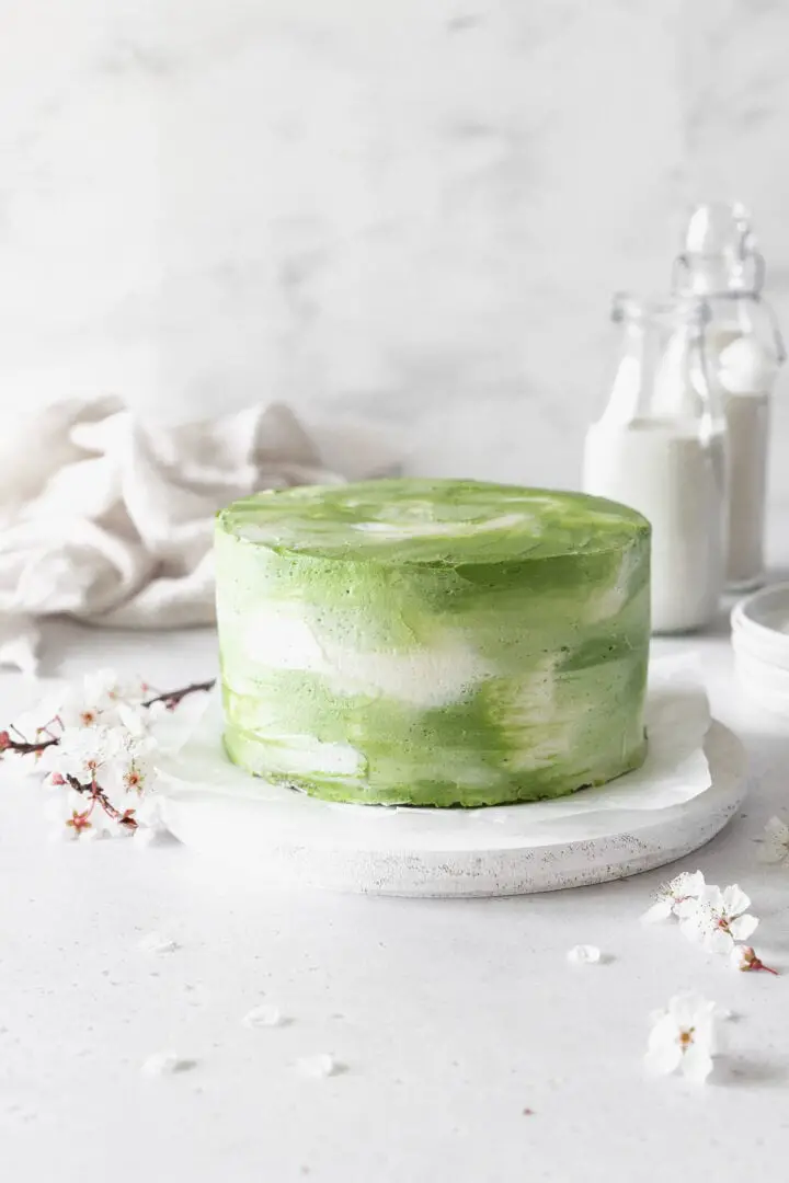 A round cake with two visible layers is covered smoothly in green and white swirled frosting, giving a soft, cloudy effect all around the sides and top. The cake sits on a piece of white parchment paper placed on a white round board, all set on a white marbled surface. Around the cake are a few delicate white flowers with dark stems, adding a natural touch. In the background, there are two clear glass containers filled with white powder and a stack of white plates, all with soft lighting and a white marbled backdrop. Photo taken with an iphone --ar 4:5 --v 7