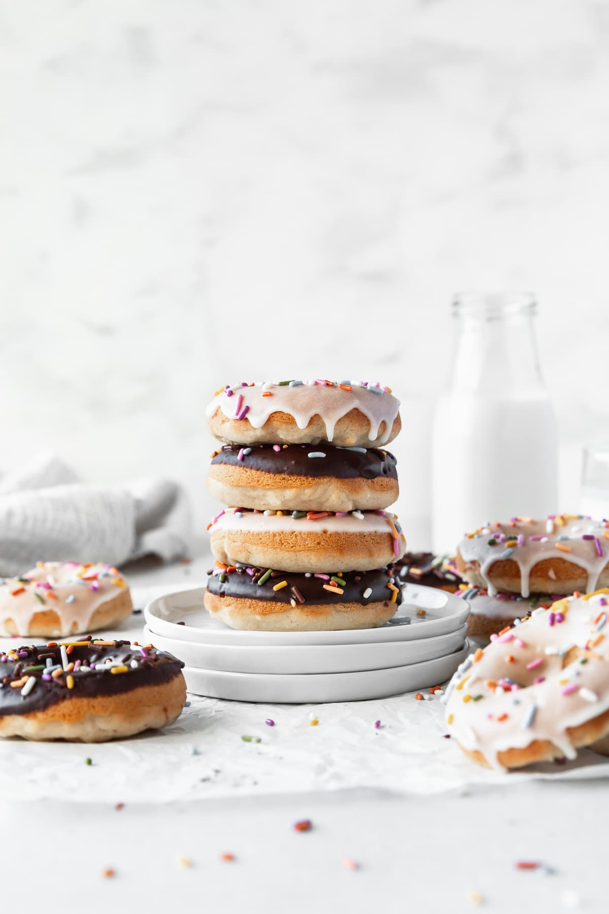 A stack of four doughnuts sits on a stack of white plates on a white marbled surface. The top doughnut has a white icing layer with colorful round and rod sprinkles, dripping slightly down the sides. The second doughnut has a smooth, dark chocolate icing with colorful rod sprinkles. The third doughnut again has white icing with sprinkles, while the bottom doughnut has chocolate icing with sprinkles. Around the plates, there are more doughnuts lying flat, some with white icing and some with chocolate icing, all sprinkled with colorful rods. In the background, there is a clear bottle of milk and a white cloth with soft folds. The whole scene is bright with a white marbled texture backdrop. photo taken with an iphone --ar 4:5 --v 7
