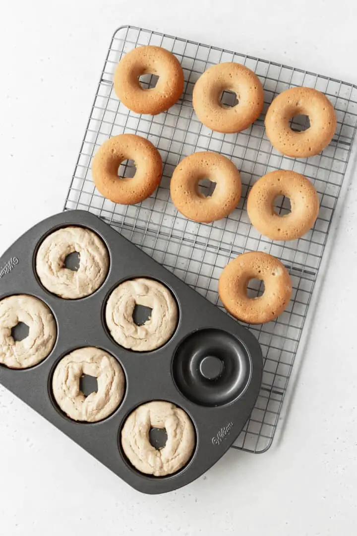 The image shows a top view of a black donut baking tray with five raw donut doughs inside the molds, each light beige with a soft and slightly cracked texture. Next to the tray is a white cooling rack holding seven fully baked donuts that are golden brown with a smooth and slightly puffy surface. The scene is set on a white marbled background, highlighting the contrast between the raw and baked donuts. Photo taken with an iphone --ar 4:5 --v 7