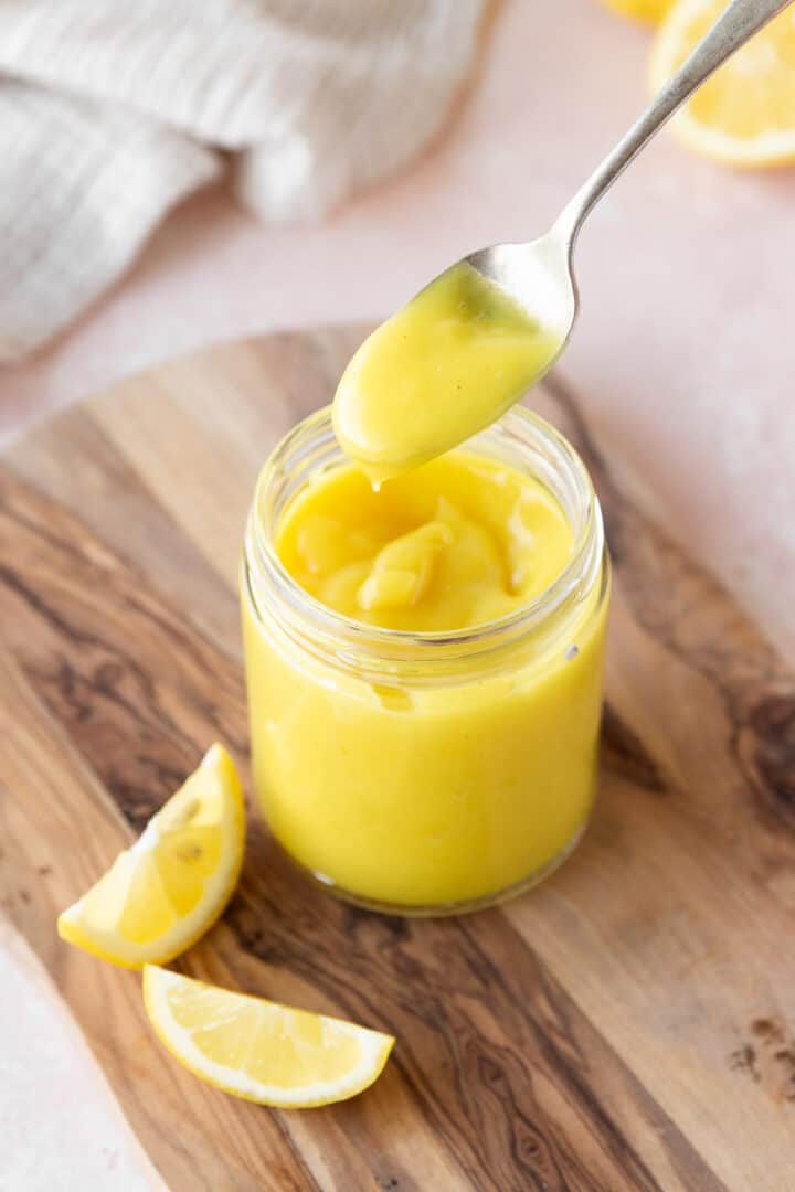 A clear glass jar filled with smooth, bright yellow lemon curd sits on a wooden board. A silver spoon holds a dollop of the thick, creamy lemon curd above the jar, showing its shiny texture. Two lemon wedges with pale yellow flesh and white rinds lie on the board near the jar, adding fresh color contrast. The scene is set on a white marbled surface with a soft beige cloth blurred in the background. photo taken with an iphone --ar 4:5 --v 7