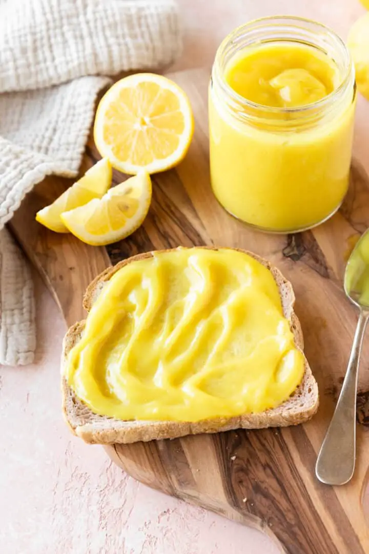 A single slice of light brown bread is spread with a thick, smooth layer of bright yellow lemon curd that has a slightly shiny texture. To the top right, there is a clear glass jar filled with more of the lemon curd showing a glossy surface with soft peaks. On the left side, there are three lemon wedges and one lemon half, all bright yellow and fresh, placed on a wooden board that holds all the items. A silver spoon with some lemon curd rests on the right edge of the board. The background is a white marbled surface with a white textured cloth near the top left corner. Photo taken with an iphone --ar 4:5 --v 7