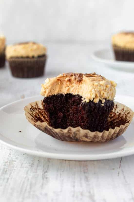 A close-up view of a chocolate cupcake with two clear layers: the bottom is a dark, rich chocolate cake with a moist texture, and the top is a thick, light caramel-colored frosting sprinkled with small chopped nuts on right side edges. The cupcake sits in a crinkled brown paper liner that is partially open, resting on a white plate placed on a white marbled surface. In the background, there are three more cupcakes that are slightly out of focus, all topped with the same caramel-colored frosting and chopped nuts. Photo taken with an iphone --ar 4:5 --v 7