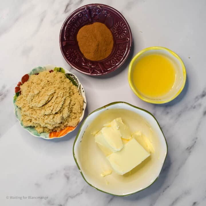 The image shows four small white bowls placed on a white marbled surface. The first bowl at the bottom right contains three pieces of pale yellow butter with a smooth texture. Above it, a bowl holds a bright yellow liquid, possibly melted butter or oil, with a shiny, smooth surface. To the left, there is a bowl full of light brown sugar, fine and crumbly in texture. At the top, another bowl contains a heap of cinnamon powder, which is deep brown and has a soft, powdery texture. All the bowls are neatly arranged to form a loose square shape. Photo taken with an iphone --ar 4:5 --v 7