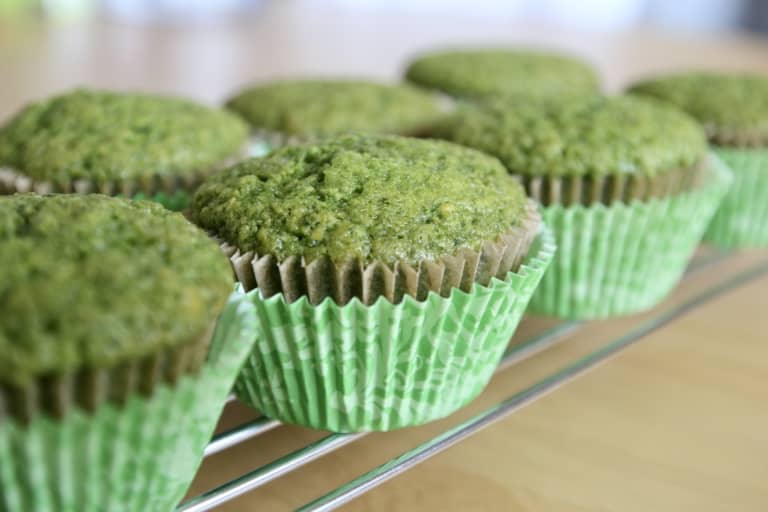 The image shows a row of green muffins in green patterned paper liners placed on a metal cooling rack. Each muffin has a slightly rough texture on top and a soft, uneven dome shape. The muffins are arranged neatly in a line, with the focus on the closest muffin while the others fade softly into the background. The cooling rack rests on a surface with a white marbled texture, and the overall lighting is natural and soft. photo taken with an iphone --ar 4:5 --v 7
