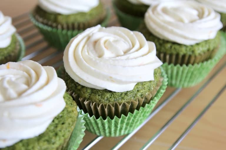 The image shows a close-up of five green cupcakes on a metal rack. Each cupcake has two layers: the bottom layer is a textured green cake with a slightly rough surface, and the top layer is a smooth, white swirl of frosting, shaped like a rose with soft, rounded edges. The cupcakes are wrapped in green paper liners with ridges. The white marbled background is barely visible behind the rack. photo taken with an iphone --ar 4:5 --v 7
