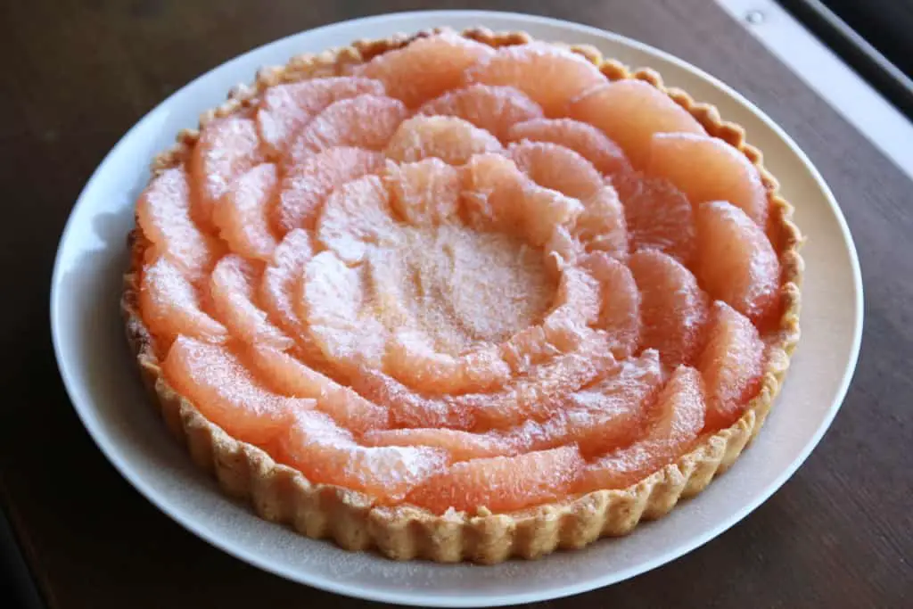 A round tart sits on a white plate with a golden-brown, slightly crumbly crust forming the base and edges. On top, there are three layers of thin, translucent pink fruit slices arranged in concentric circles starting from the outer edge moving inward, each layer slightly overlapping the one below. The topmost fruit layer is dusted lightly with white powdered sugar, giving it a soft, frosted look. The whole tart is presented on a white marbled surface. Photo taken with an iphone --ar 4:5 --v 7