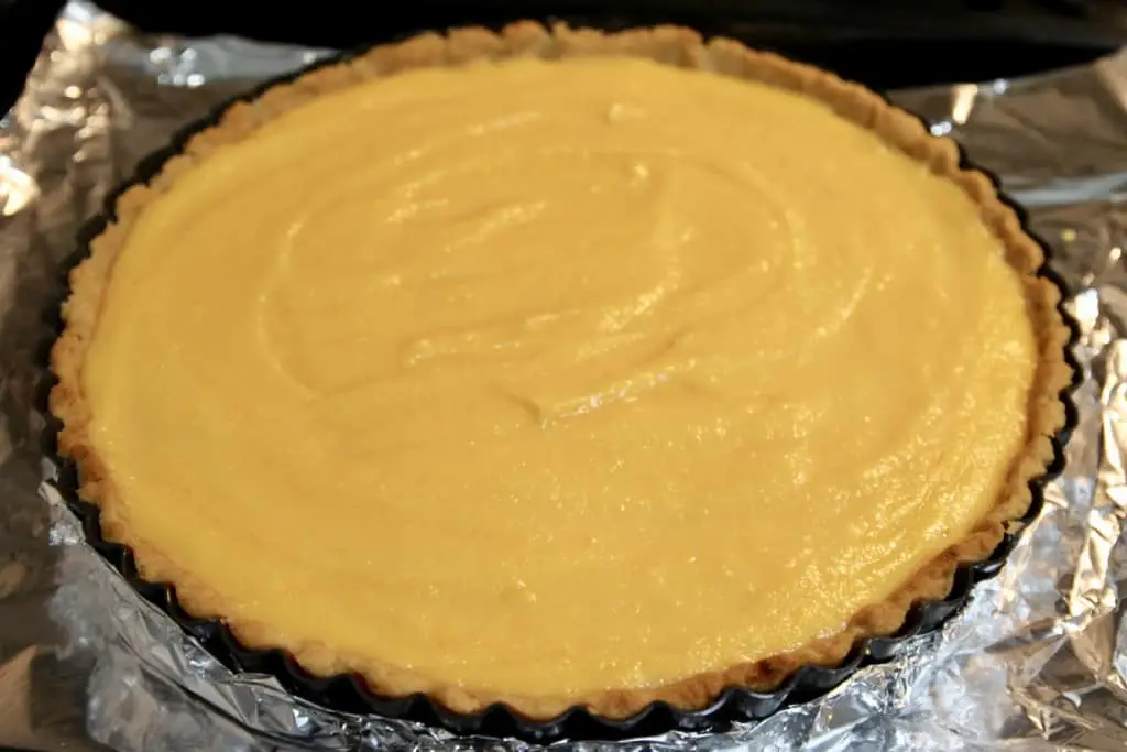 The image shows a tart with one layer, featuring a smooth pale yellow filling spread evenly in a round tart shell with a light golden brown crust that has a slightly scalloped edge. The tart sits on a piece of shiny aluminum foil against a dark background. photo taken with an iphone --ar 4:5 --v 7