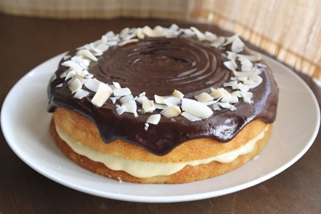 A two-layer round cake on a white plate, each layer is golden brown with a thick middle layer of smooth light cream filling visible between them. The top layer is covered with dark, glossy chocolate frosting that slightly drips over the cake sides. There is a circle of white coconut flakes on top edges, contrasting with the dark chocolate. The plate sits on a white marbled surface. photo taken with an iphone --ar 4:5 --v 7