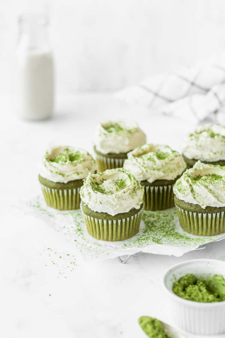 There are six green cupcakes arranged on a white surface with a white doily underneath some of them. Each cupcake has a single swirl of white frosting on top, sprinkled with green powder. In the foreground, there is a white bowl filled with green powder and a spoon with some green powder on it. The background shows a white bottle and a white cloth with a black grid pattern on a white marbled surface. Photo taken with an iphone --ar 4:5 --v 7