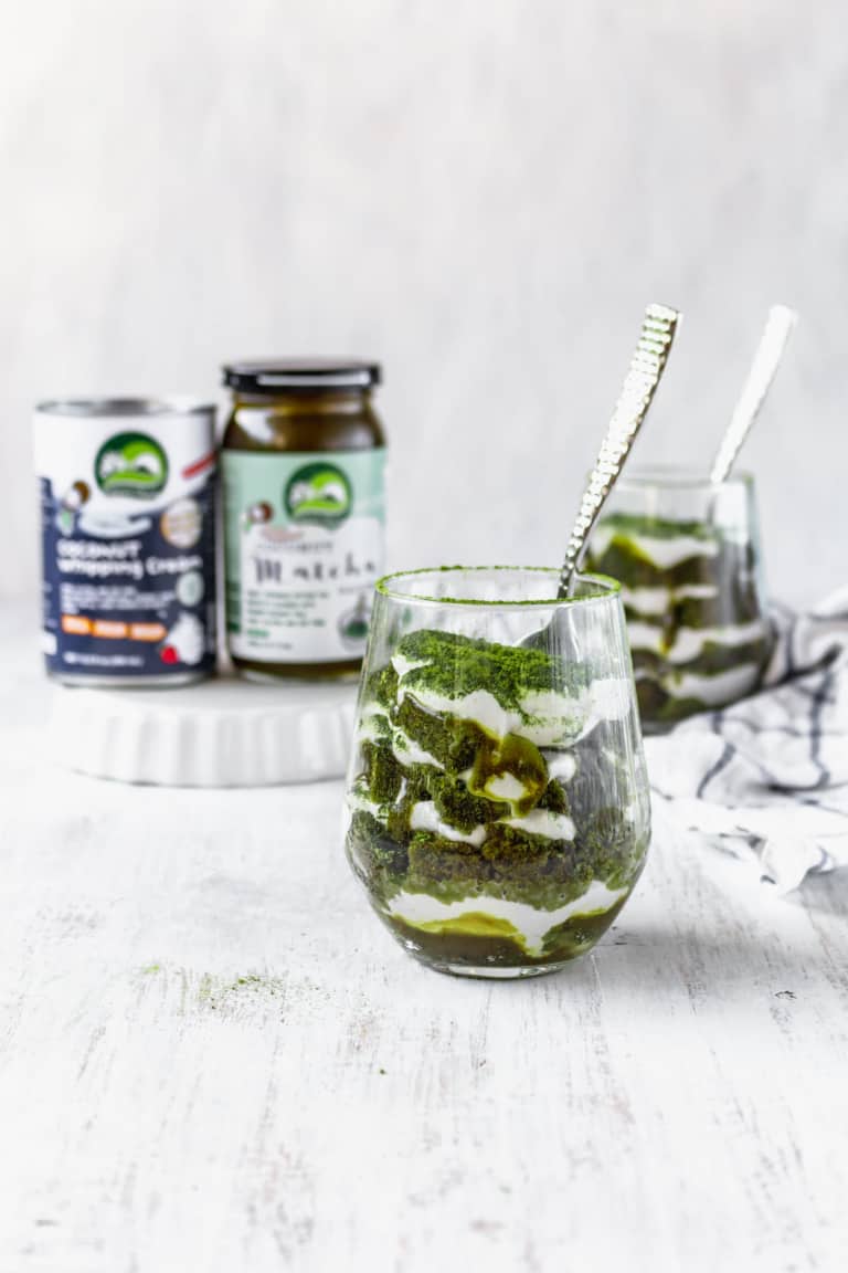 The image shows two clear glasses filled with a layered dessert placed on a white marbled surface. Each glass has alternating layers of dark green matcha powder and white creamy yogurt or cream, with about five visible layers in each glass. The dark green layers have a powdery texture, while the white layers are smooth and creamy. One glass is in the front center with a silver spoon standing inside, and the second glass is slightly blurred in the background to the right, also with a silver spoon. Behind the glasses, two jars with green and white labels are placed on a small white plate, slightly out of focus, adding depth to the image. The overall setting is bright with a clean and soft light. Photo taken with an iphone --ar 4:5 --v 7