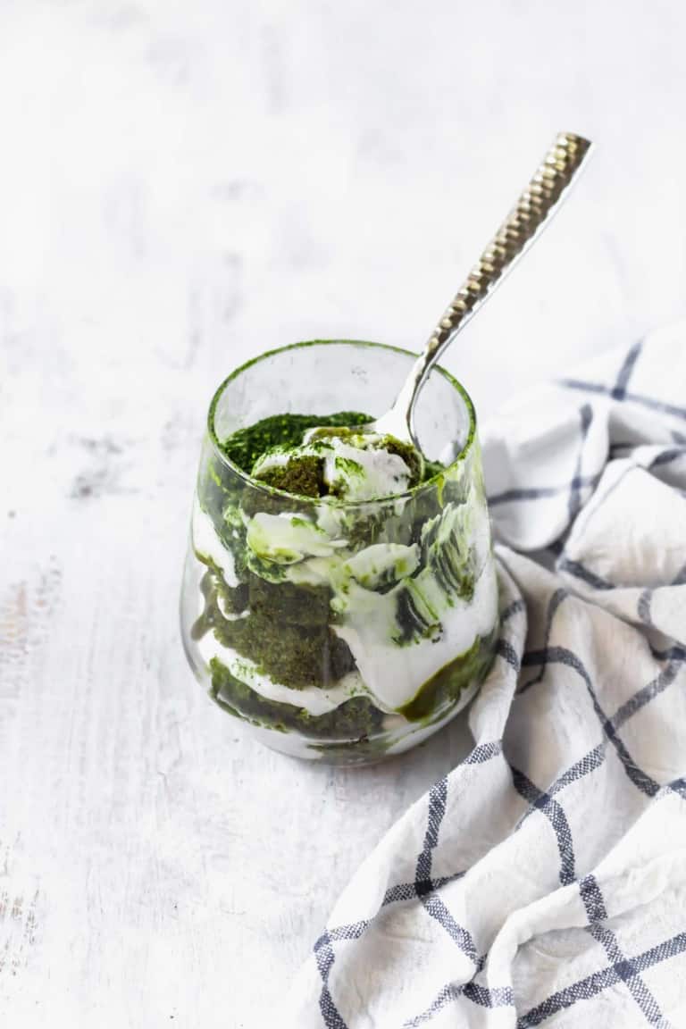 A clear glass holds a layered dessert with three visible layers, starting with a dark green textured base, followed by a smooth white creamy layer, then another thick dark green layer with a slightly rough texture. The white cream is swirled unevenly throughout the green layers, creating a marbled effect. A silver spoon with a hammered handle is placed inside the glass, scooping from the top. The glass sits on a white marbled surface next to a white cloth with black grid lines. photo taken with an iphone --ar 4:5 --v 7