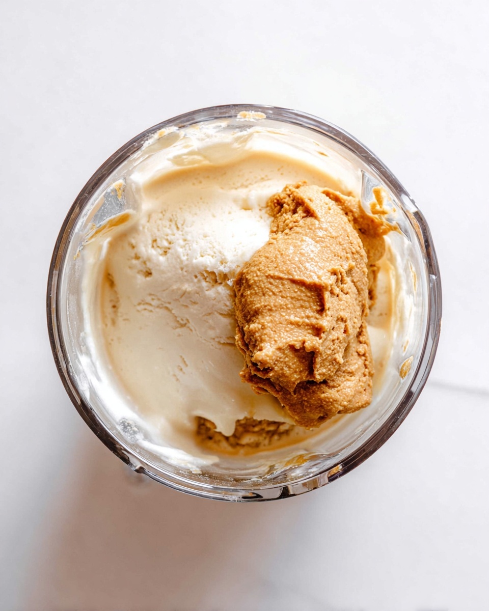A clear blender cup is shown from above with two main layers inside: a creamy off-white ice cream layer that looks soft and smooth at the bottom, topped with a thick scoop of light brown peanut butter that has a slightly rough and textured surface. The blender cup is set against a white marbled background. photo taken with an iphone --ar 4:5 --v 7