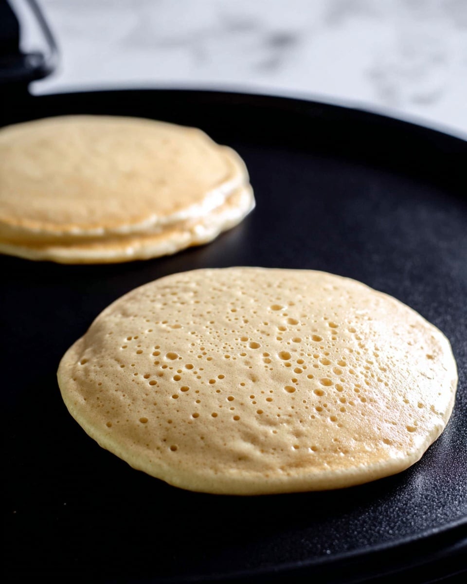 Pancakes for One Recipe 5 The image shows two round, thick pancake batter portions cooking on a black griddle. The pancakes have a light beige color with bubbles appearing on the surface, indicating they are in the process of cooking. The texture looks smooth but slightly uneven, with soft edges starting to firm up. The background is a white marbled surface, and some black cords from the griddle are visible in the upper part of the image. photo taken with an iphone --ar 4:5 --v 7