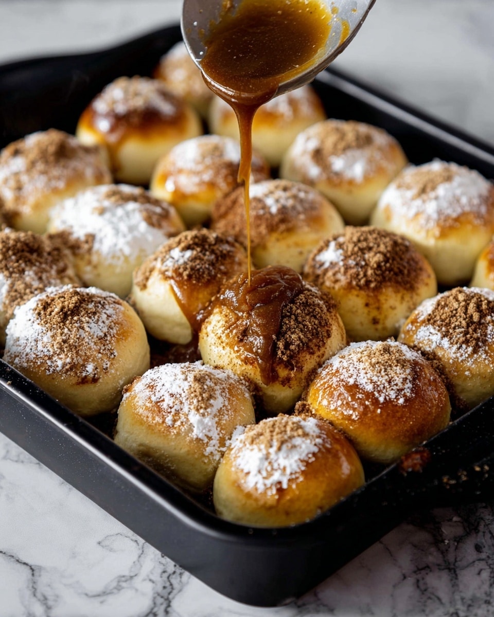 The image shows a black tray filled with round bread rolls, each topped with a dusting of white powder and a sprinkling of brown spice. A spoon is pouring a thick, dark caramel-colored sauce over one roll in the front right corner. The rolls are arranged closely in the tray, showing a soft, golden-brown surface under the toppings. The tray is placed on a white marbled surface. photo taken with an iphone --ar 4:5 --v 7
