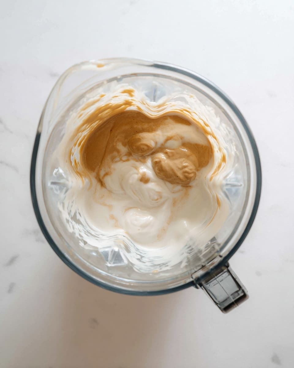 A clear glass blender cup is shown from above, sitting on a white marbled surface. Inside the blender, there are two main layers: the bottom layer is smooth and creamy white, while the top layer is a light tan liquid poured over the white layer, creating soft swirls where they mix. The texture of the top layer looks slightly thick and uneven. The blender cup has a handle on the upper right side, and the light reflects softly on the glass. photo taken with an iphone --ar 4:5 --v 7