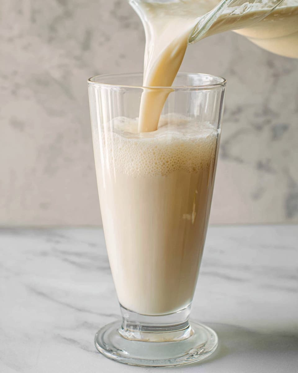 A tall clear glass with a thick, creamy light beige liquid being poured into it from a clear glass pitcher positioned at the top right, the liquid creating soft frothy bubbles on the surface inside the glass. The glass is placed on a white marbled surface with subtle gray veins, and the background is also a soft white marbled texture, creating a clean and minimal look. The light softly highlights the smooth texture of the liquid and glass. photo taken with an iphone --ar 4:5 --v 7