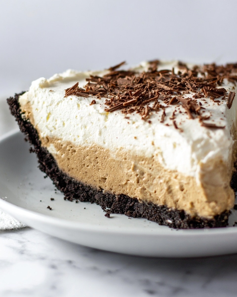 A slice of pie on a white plate shows three layers: the bottom layer is a dark crumbly base made from crushed cookies, the middle layer is thick and light brown with a smooth and creamy texture, and the top layer is fluffy white cream sprinkled with thin dark chocolate shavings. The pie is positioned on a white marbled surface, and the focus is close up, capturing the pie’s creamy and soft textures clearly. photo taken with an iphone --ar 4:5 --v 7