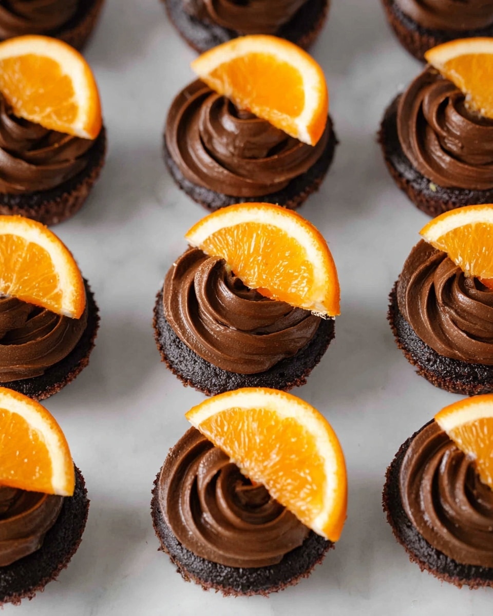 The image shows a group of small chocolate cupcakes arranged in rows on a white marbled surface. Each cupcake has two clear layers: a dark chocolate cake base and a thick, smooth swirl of dark chocolate frosting on top. Sitting on the frosting of each cupcake is a bright, shiny orange slice placed off-center, adding a fresh pop of color against the deep brown. The cupcakes are evenly spaced, and the texture of the frosting looks creamy and soft, contrasting with the denser cake layer below. photo taken with an iphone --ar 4:5 --v 7