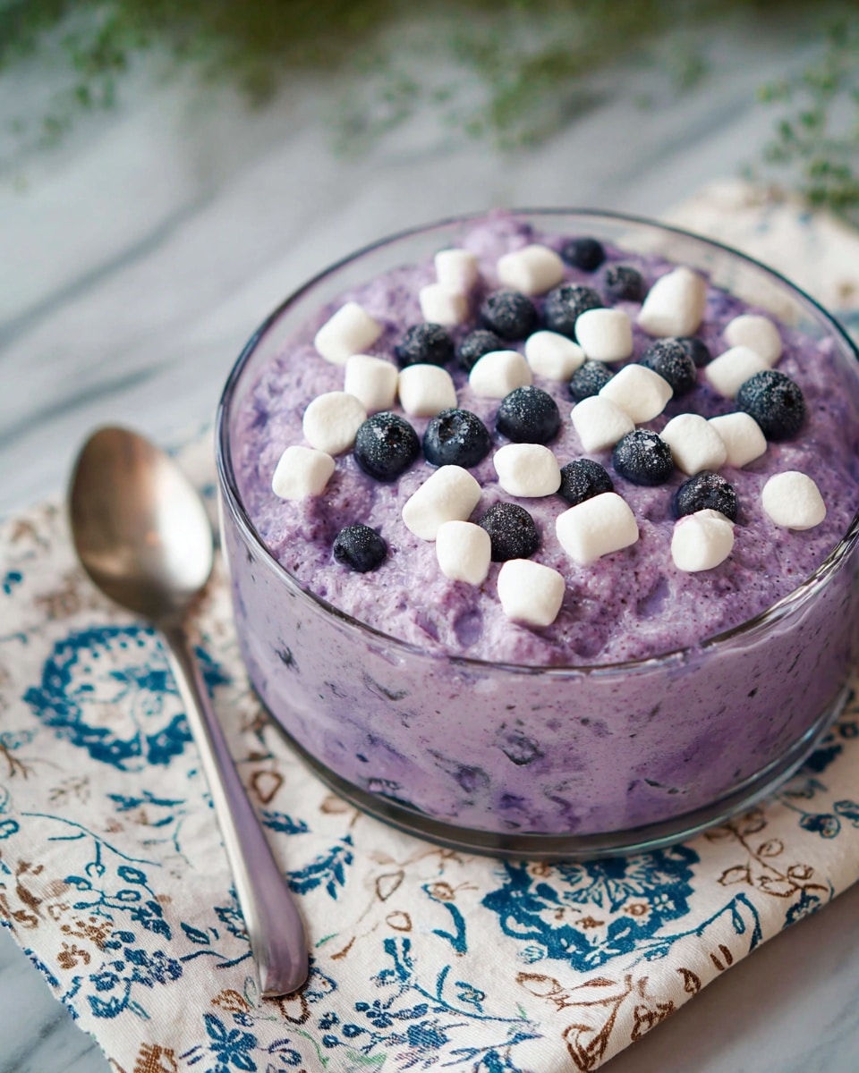 A clear glass bowl filled with a thick purple mixture that has visible small dark pieces inside it, topped with small white mini marshmallows and scattered dark blueberries on the surface, sitting on a white cloth with blue and brown floral patterns, all on a white marbled surface. Nearby is a metal spoon placed on the cloth in the front left corner of the image, photo taken with an iphone --ar 4:5 --v 7