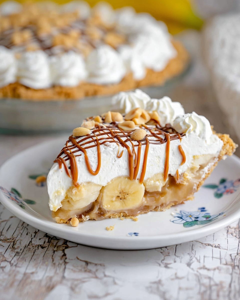 A slice of pie on a white plate with a small blue flower pattern sits on a white marbled surface. The slice has a bottom layer of golden crust, followed by a thick layer of banana pieces and light brown filling. On top is a thick, white whipped cream layer decorated with a crisscross pattern of thin caramel and chocolate drizzle and scattered peanuts. The edges of the pie slice have additional piped white cream swirls. In the background, a full pie on a white plate with white whipped cream swirls around the edge is partly visible. Photo taken with an iphone --ar 4:5 --v 7