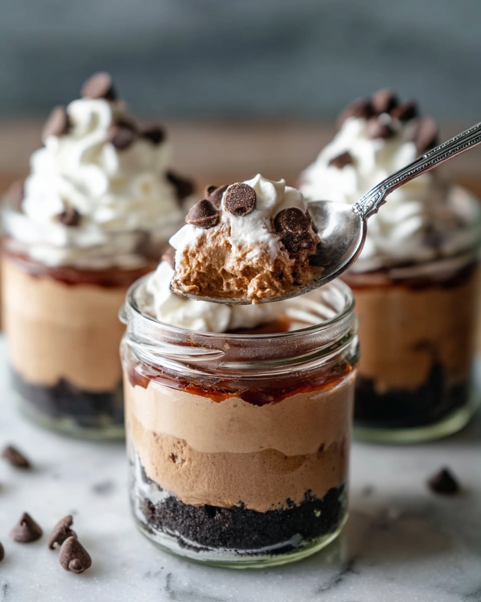 Three small glass jars hold a layered dessert on a white marbled surface. Each jar has three layers: at the bottom, a dark crumbly base; the middle layer is thick and light brown with a smooth texture; the top layer is a dollop of white whipped cream with small dark chocolate chips scattered on it. One jar is in the front with a silver spoon scooping from the top layer, showing the creamy texture of the dessert, while the other two jars are blurred in the background. The lighting is soft and natural, highlighting the creamy and crunchy textures. Photo taken with an iphone --ar 4:5 --v 7