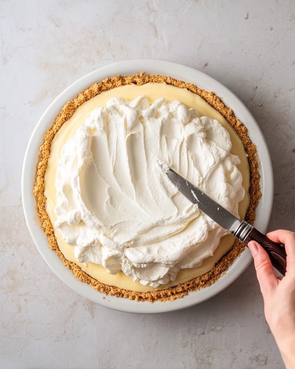 A white plate holds a pie with three layers visible: the bottom is a thick, crimped golden brown crust with a textured, slightly rough surface; above it is a smooth, pale yellow cream filling spread evenly just inside the crust edges; the top layer is a thick, fluffy white whipped topping being spread by a woman's hand holding a small knife with a dark wooden handle and a shiny metal blade. The scene is set on a white marbled surface. Photo taken with an iphone --ar 4:5 --v 7