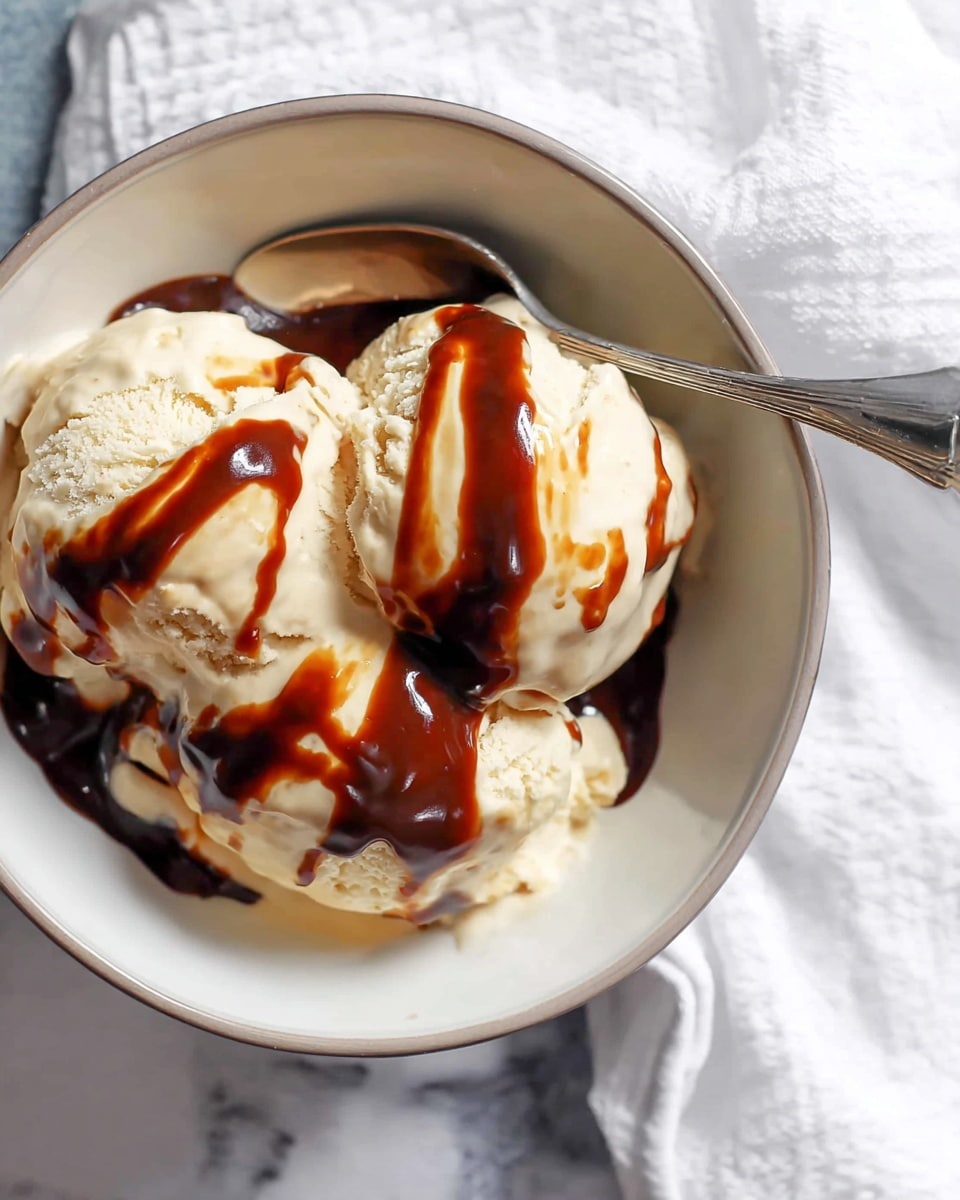A white bowl filled with three large scoops of light beige ice cream with a soft, creamy texture. The ice cream is topped with a thick, glossy dark brown sauce drizzled unevenly across the scoops. A silver spoon rests on the edge of the bowl, partially touching the ice cream. The bowl sits on a white marbled surface with a white cloth nearby. photo taken with an iphone --ar 4:5 --v 7