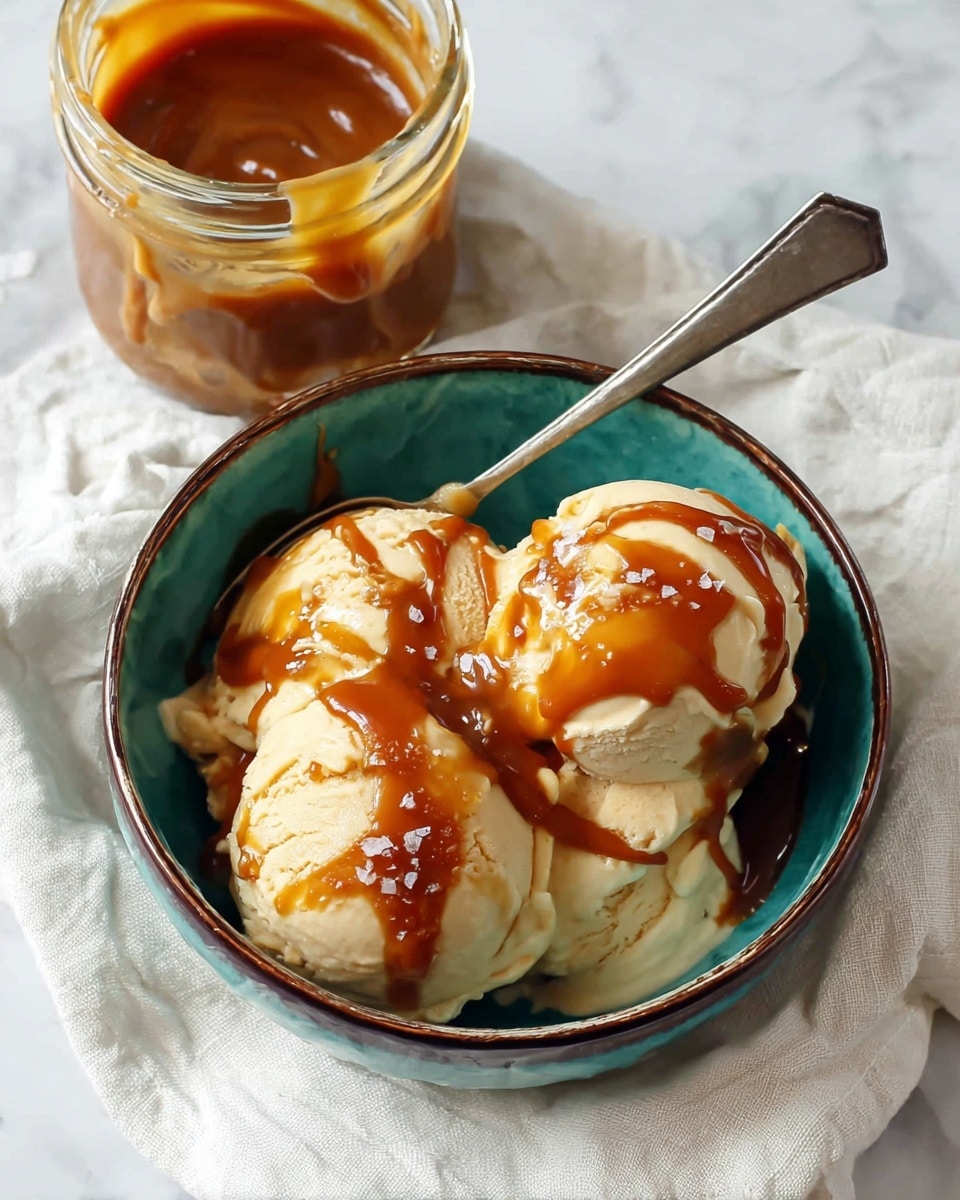 A turquoise bowl with a dark rim holds three large, smooth, cream-colored scoops of ice cream, each generously drizzled with thick, dark caramel sauce on top, creating rich swirls and patches. The sauce shines slightly, with a few flaky bits of salt scattered on it. A silver spoon rests inside the bowl, its handle extending out over the edge. To the left of the bowl, there is a partially open jar filled with the same caramel sauce, showing its creamy texture inside. All of this is placed on a soft, white cloth, set upon a white marbled surface. Photo taken with an iphone --ar 4:5 --v 7