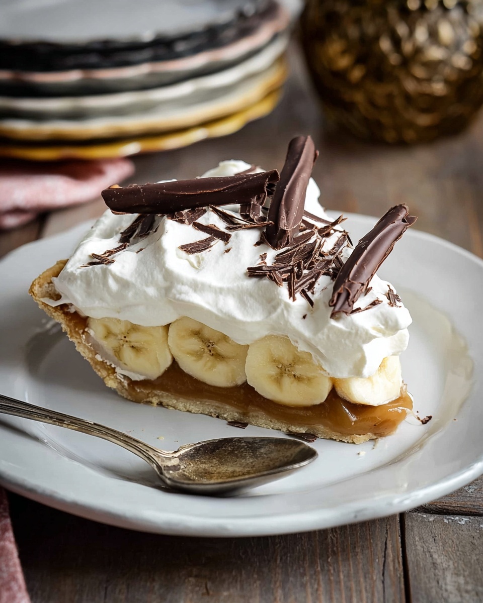 A slice of pie is shown on a white plate with a vintage silver spoon beside it. The pie has four visible layers: a golden-brown crust at the bottom, a caramel-colored filling, a layer of sliced bananas that are pale yellow, and a thick layer of white whipped cream on top. The whipped cream is decorated with several dark brown chocolate shavings and two longer chocolate sticks placed diagonally. The setting includes a rustic wooden table and blurred background with plates stacked and a decorative object. The photo taken with an iphone --ar 4:5 --v 7
