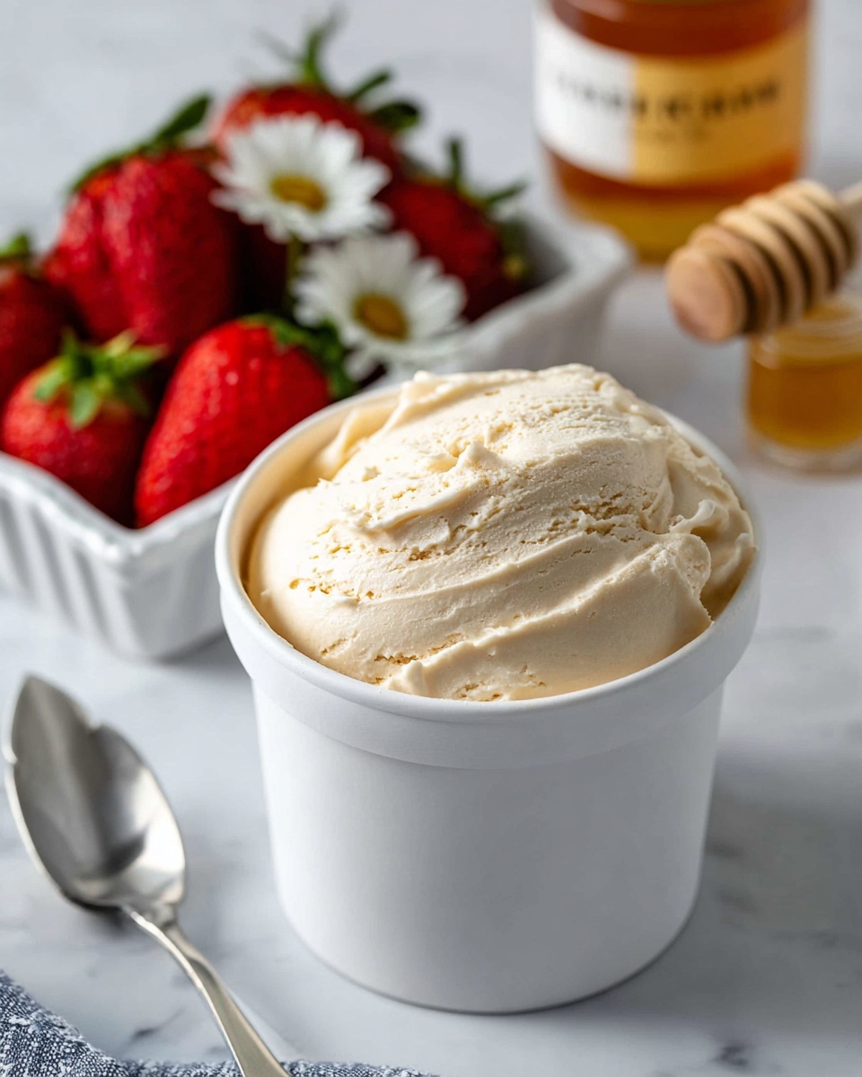 The image shows a white cylindrical container filled with smooth, creamy ice cream that has a pale beige color on top with soft swirls and gentle peaks. The container sits on a white marbled surface, and there is a white scalloped bowl with a silver spoon on the left side. In the background, a white square bowl holds bright red strawberries with green tops and a small white flower. To the right, there is a small jar with golden honey and a golden honey dipper nearby. The whole scene is bright and fresh, focusing on the creamy texture of the ice cream and the vivid red of the strawberries, photo taken with an iphone --ar 4:5 --v 7
