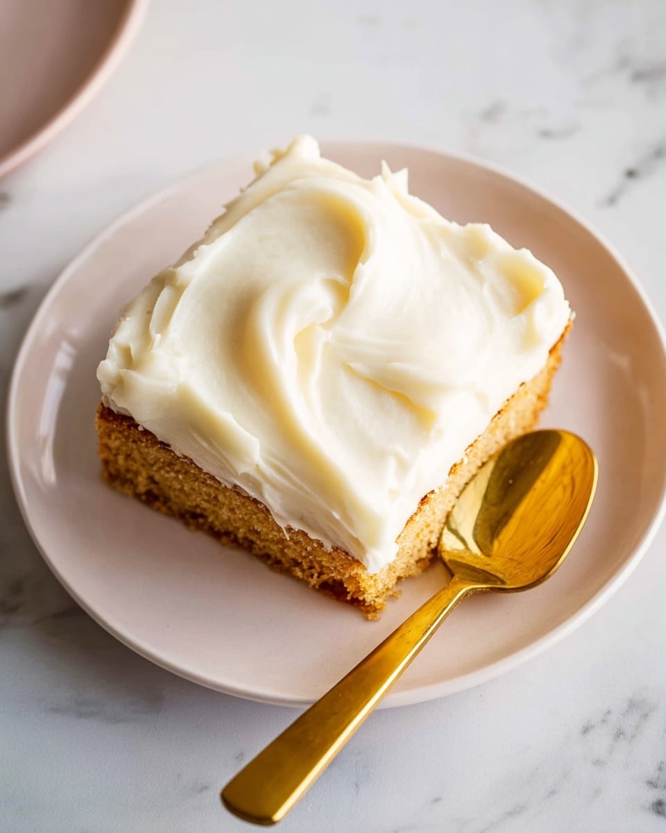 A single square piece of cake with a light golden brown base layer is topped with a thick, creamy layer of smooth white frosting that has soft, swirled textures covering the entire top. The cake sits on a simple white plate, and next to it is a shiny gold spoon resting on the plate's right side. The setting is on a white marbled surface, adding subtle texture to the background. Photo taken with an iphone --ar 4:5 --v 7