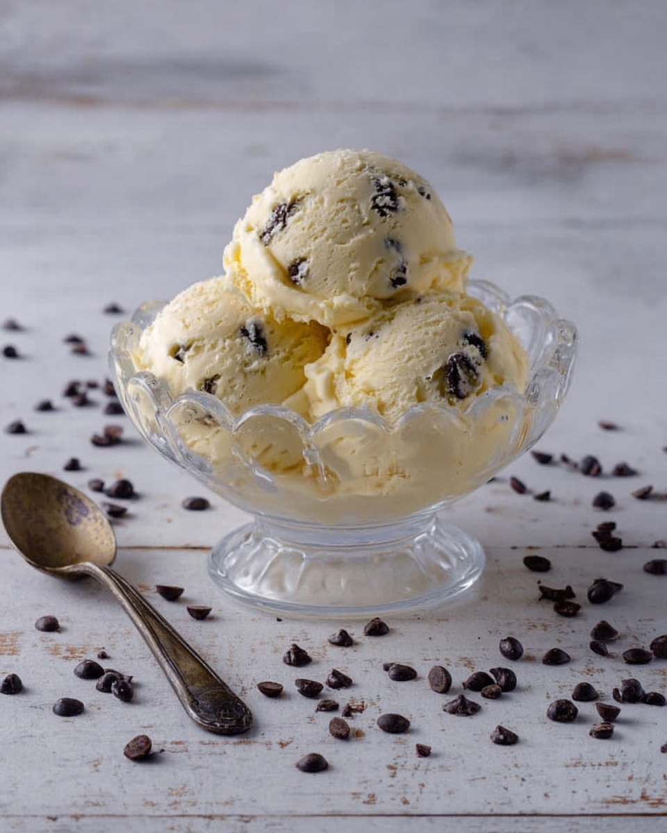 A clear glass bowl with scalloped edges holds three scoops of pale yellow ice cream with dark chocolate chips mixed in. The bowl sits on a white marbled surface scattered with many small, dark chocolate chips around it. To the left of the bowl, there is a tarnished silver spoon resting on the surface. The ice cream texture looks smooth and creamy with bits of chocolate visible throughout. The photo taken with an iphone --ar 4:5 --v 7