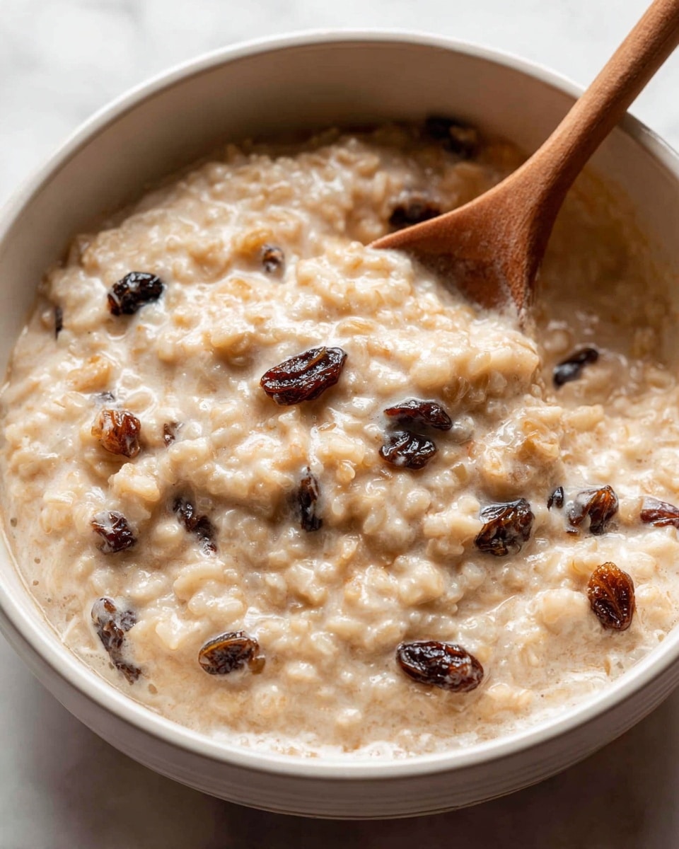 A close-up of a hot creamy rice pudding in a white bowl with a thick texture, showing soft rice grains mixed with dark raisins spread throughout. The pudding has a light beige color with a smooth and slightly grainy surface. A wooden spoon is dipped into the pudding on the right side, mixing the contents gently. The bowl sits on a white marbled surface with soft natural light highlighting the pudding's creamy and tender texture photo taken with an iphone --ar 4:5 --v 7