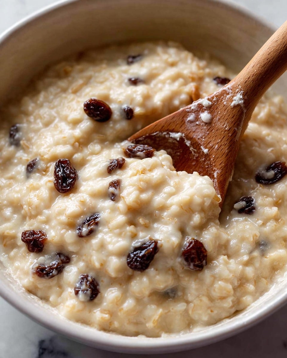 A close-up view of a creamy rice pudding with visible soft rice grains mixed with plump dark raisins scattered throughout. It is served in a white bowl that shows some rice pudding smeared on the inner rim. A wooden spoon is partially submerged in the pudding, showing the thick and smooth texture. The background is a white marbled texture. photo taken with an iphone --ar 4:5 --v 7
