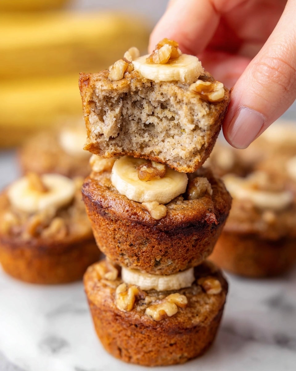 A stack of four small banana muffins sits on a white marbled surface, each muffin showing a golden-brown crust with visible small walnut pieces on top. The top muffin is held by a woman's hand, broken in half to reveal its soft, moist interior speckled with banana bits. Each muffin has a thin banana slice on top beneath the walnuts, with a slightly cracked texture on the surface and a spongy texture inside. The background is blurred with more banana muffins and bananas visible, enhancing the warm and fresh feel of the image. Photo taken with an iphone --ar 4:5 --v 7