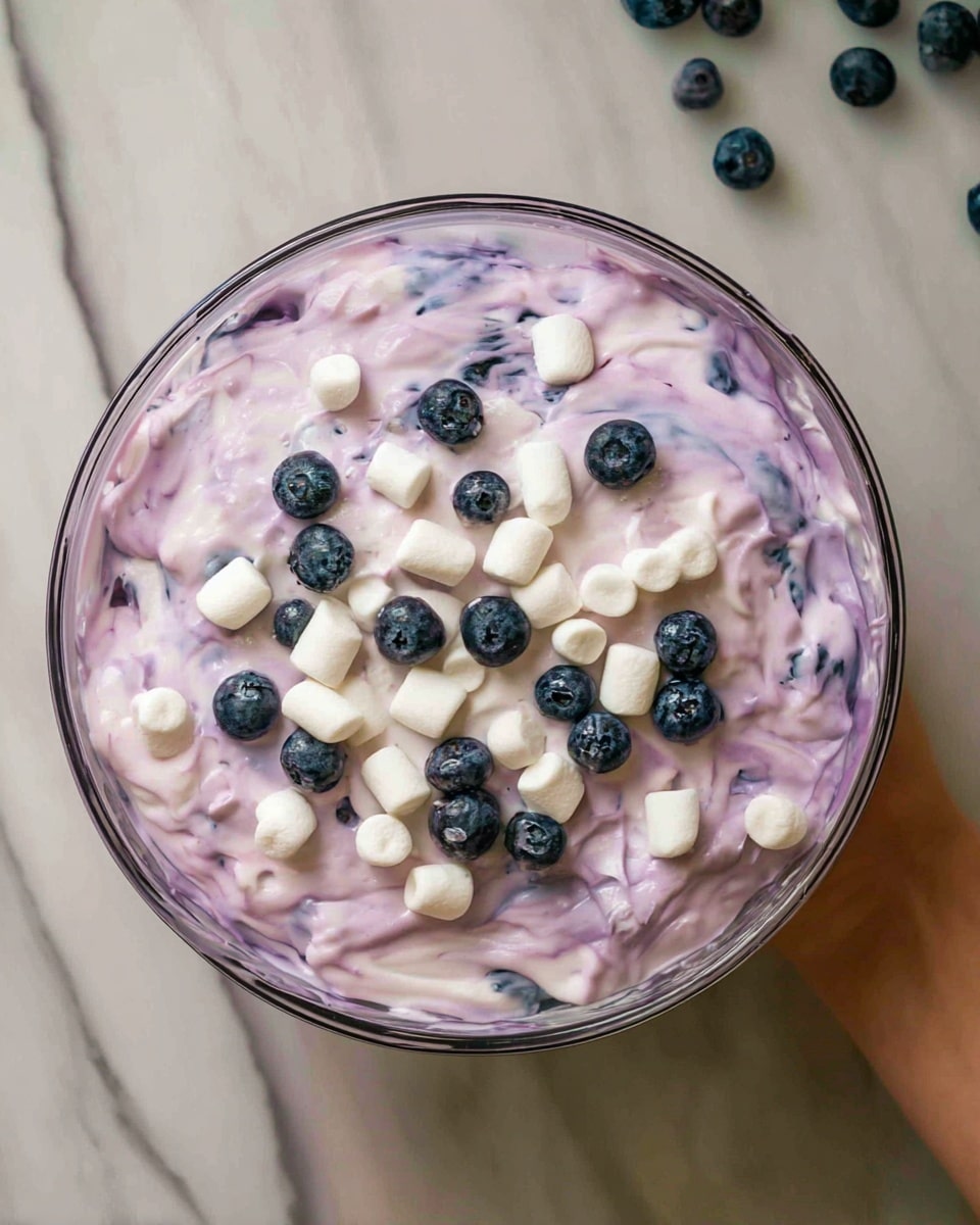 A clear glass bowl filled with a creamy mixture of light purple and white swirled together, creating a smooth and soft texture. On top, there are scattered small white marshmallows and fresh dark blue blueberries, adding round pops of color. The bowl sits on a white marbled surface, and part of a woman's hand is visible holding the edge of the bowl. photo taken with an iphone --ar 4:5 --v 7
