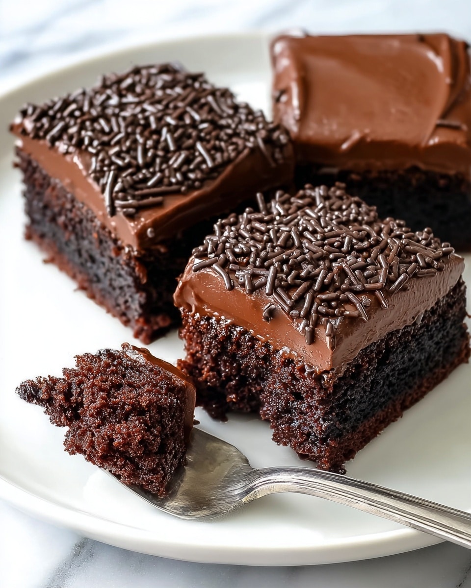 There are three square pieces of chocolate cake on a white plate. Each piece has two layers: a dark brown, moist cake base and a thick, smooth chocolate frosting on top. The front piece is decorated with chocolate sprinkles covering the frosting. A silver spoon holds a bite-sized piece from the front square, showing the soft cake and frosting layers inside. The plate sits on a white marbled surface. Photo taken with an iphone --ar 4:5 --v 7