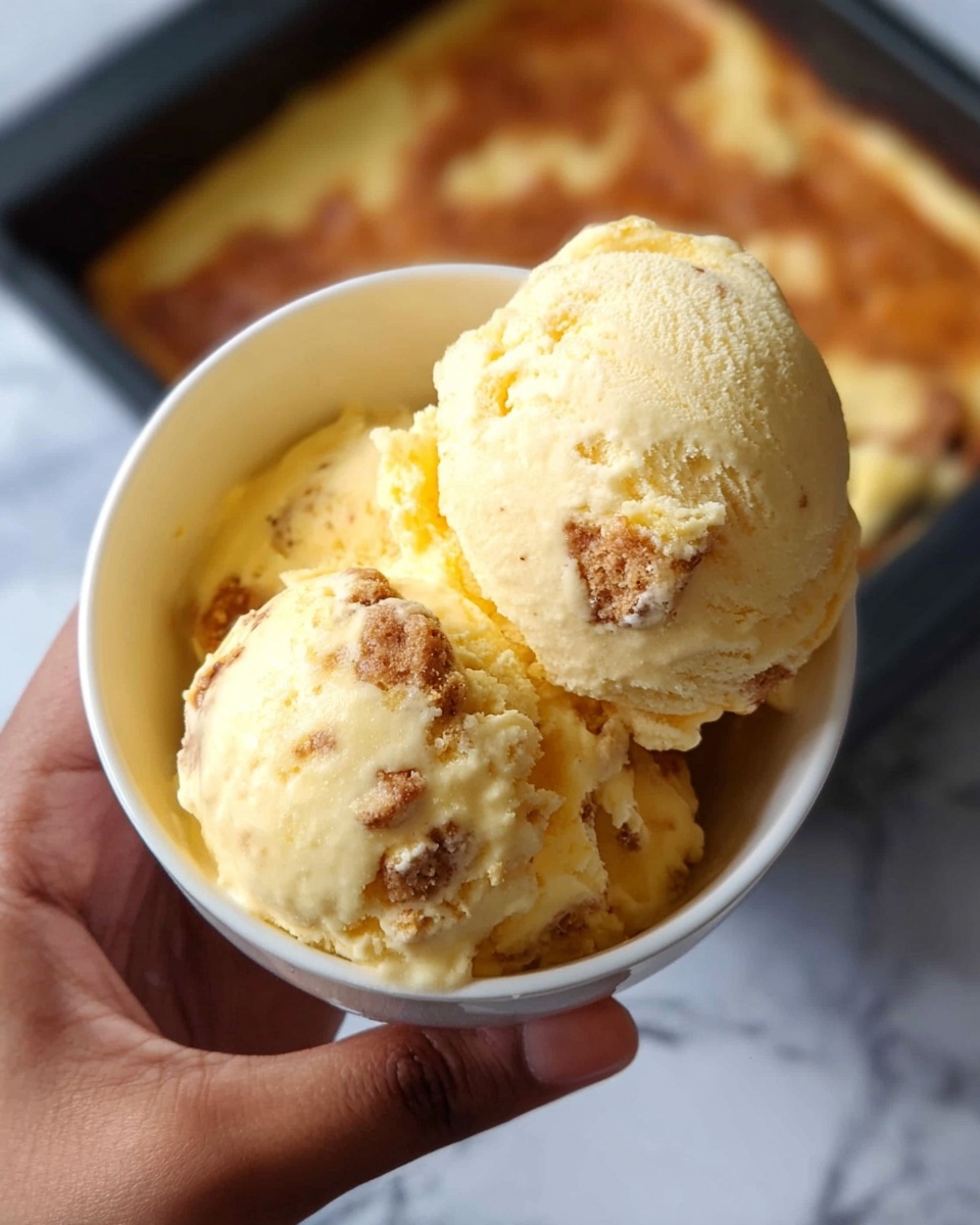 A white bowl holds two scoops of light yellow ice cream with visible brown crumbs mixed in. The ice cream texture looks smooth but slightly cracked on the surface. The bowl is lifted by a woman’s hand with a medium skin tone, showcasing a close-up view. In the background, a baking tray with a flat layer of browned baked dessert contrasts with a white marbled surface beneath. Photo taken with an iphone --ar 4:5 --v 7