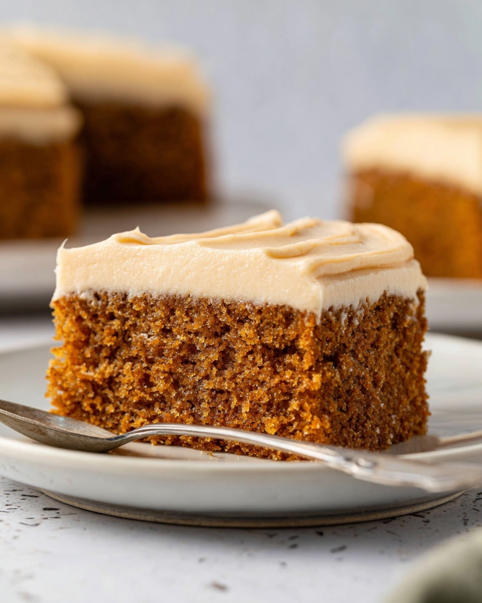 Pumpkin Olive Oil Cake with Cream Cheese Frosting Recipe 6 The image shows a close-up of a single square slice of cake on a white plate with a spoon beside it. The cake has two layers: the bottom layer is thick, moist, and brown with a crumbly texture, and the top layer is a creamy, light beige frosting spread evenly with a smooth and soft appearance. In the background, there are two more slices of the same cake, slightly out of focus, set on a white marbled surface. The photo was taken with an iphone --ar 4:5 --v 7