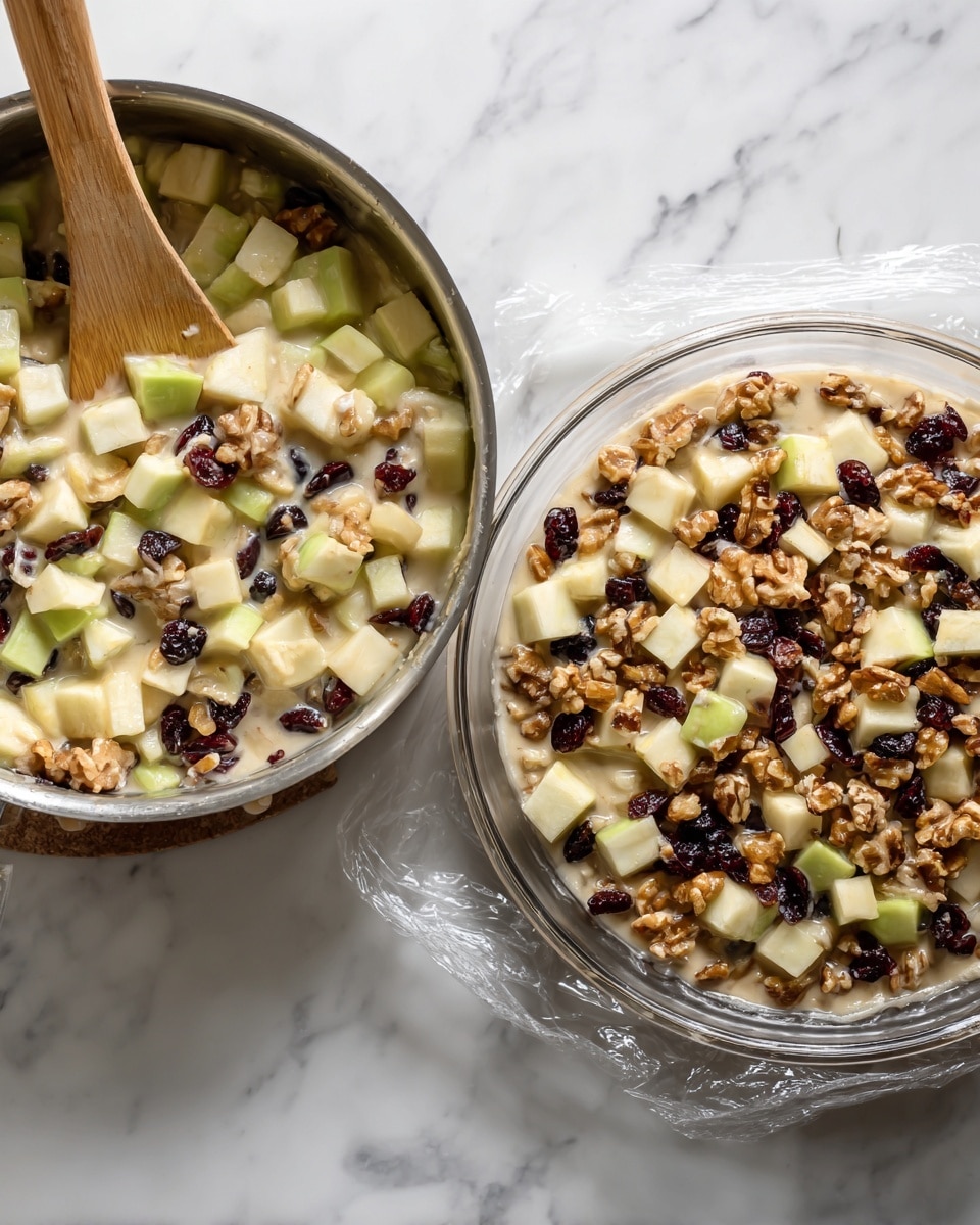 The image shows a two-part scene: on the left is a close-up of a metal pan filled with mixed ingredients—light green cubed apple pieces, dark red dried cranberries, and whole walnut pieces—held together with a thick pale sauce, with a wooden spoon resting inside the pan. On the right is a clear glass pie dish sitting on a white marbled surface, filled evenly to the top with the same mixture of pale apple cubes, dark dried cranberries, and tan walnuts coated in the creamy sauce. A piece of clear plastic wrap is loosely covering the pie dish. photo taken with an iphone --ar 4:5 --v 7