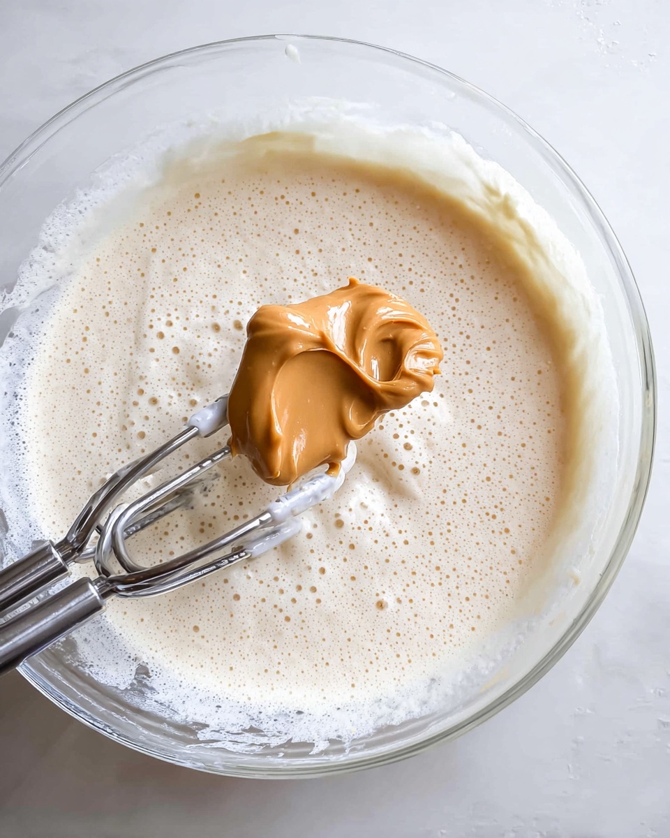 The image shows a close-up of a clear glass bowl filled about three-quarters with a frothy, light cream mixture that has a smooth and airy texture with small bubbles on the surface. Inside the bowl, there is a metallic electric mixer with two beaters resting on the left side, partially immersed in the mixture. In the center, a silver spoon is held above the mixture, carrying a dollop of thick, smooth, golden-brown peanut butter with a slightly shiny surface. The scene is set on a white marbled texture background, creating a clean and bright environment. photo taken with an iphone --ar 4:5 --v 7