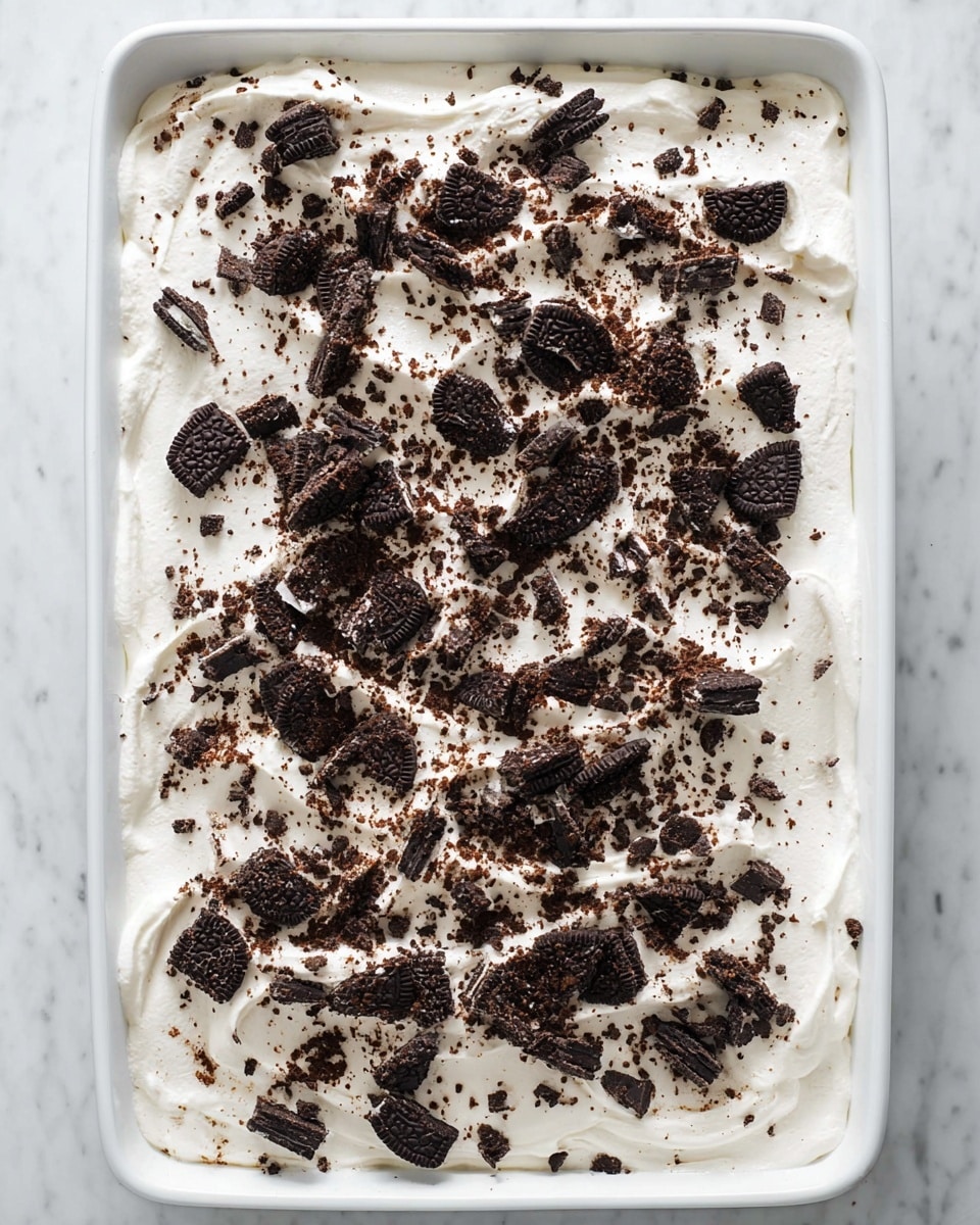 A rectangular white dish filled with a thick, even layer of white whipped cream topped with many small and large pieces of dark chocolate sandwich cookies scattered all over. The cookies vary in size, with some crushed into fine crumbs and others in bigger chunks, creating a contrast with the smooth cream beneath. The dish sits on a white marbled surface, and the overall look is fresh and inviting. photo taken with an iphone --ar 4:5 --v 7