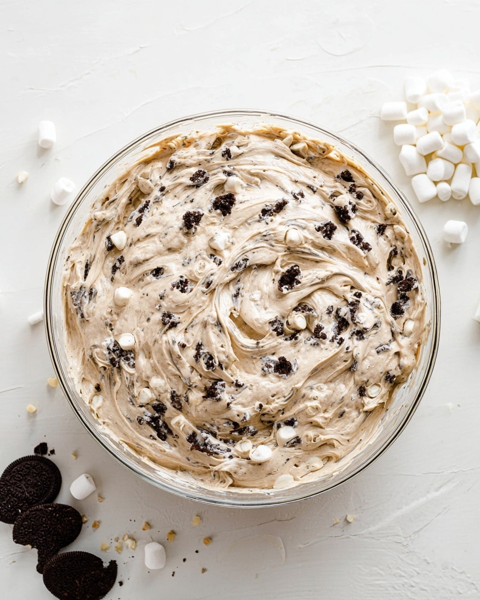 A clear glass bowl filled with a thick, creamy mixture that is light brown with visible small white chunks and dark cookie pieces blended evenly throughout, creating a textured swirled surface on top. The bowl sits on a white marbled background with some small white marshmallows gathered on the right side and broken dark chocolate cookies scattered near the bottom left corner. The overall look is soft and fluffy with a mix of creamy and crunchy bits photo taken with an iphone --ar 4:5 --v 7